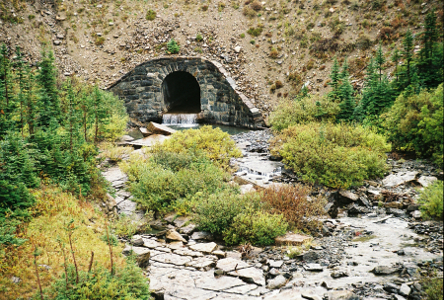 [A stone arch surrounding an opening on the side of a hill. There is water flowing thorough the arch and into the foreground over what appear to be broken stones.]